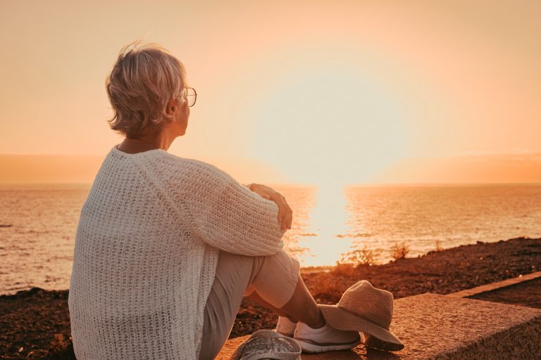 Back view of mature woman sitting alone face the sea at sunset light looking at the horizon.