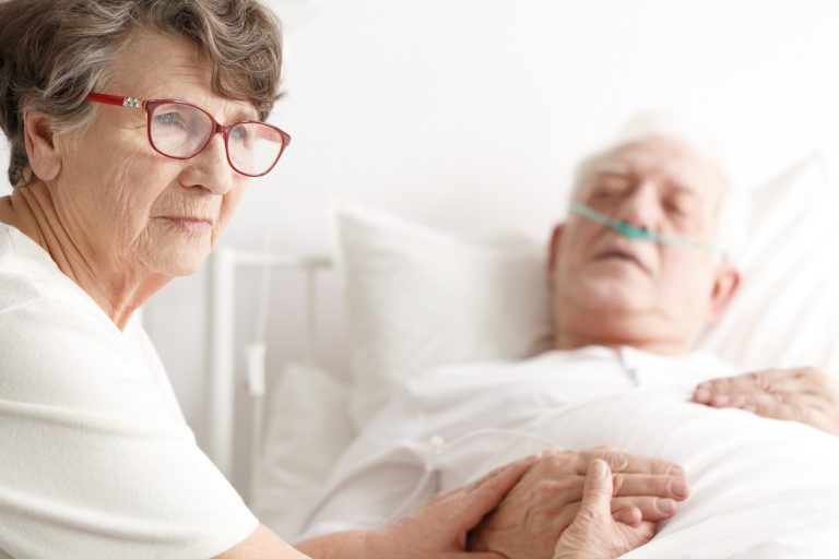 Sad senior woman caregiver holding hands with husband in bed