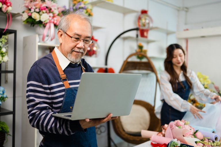 senior man florist talking on smartphone using laptop at florist. portrait of mature male small business owner using laptop and looking at camera in flower shop