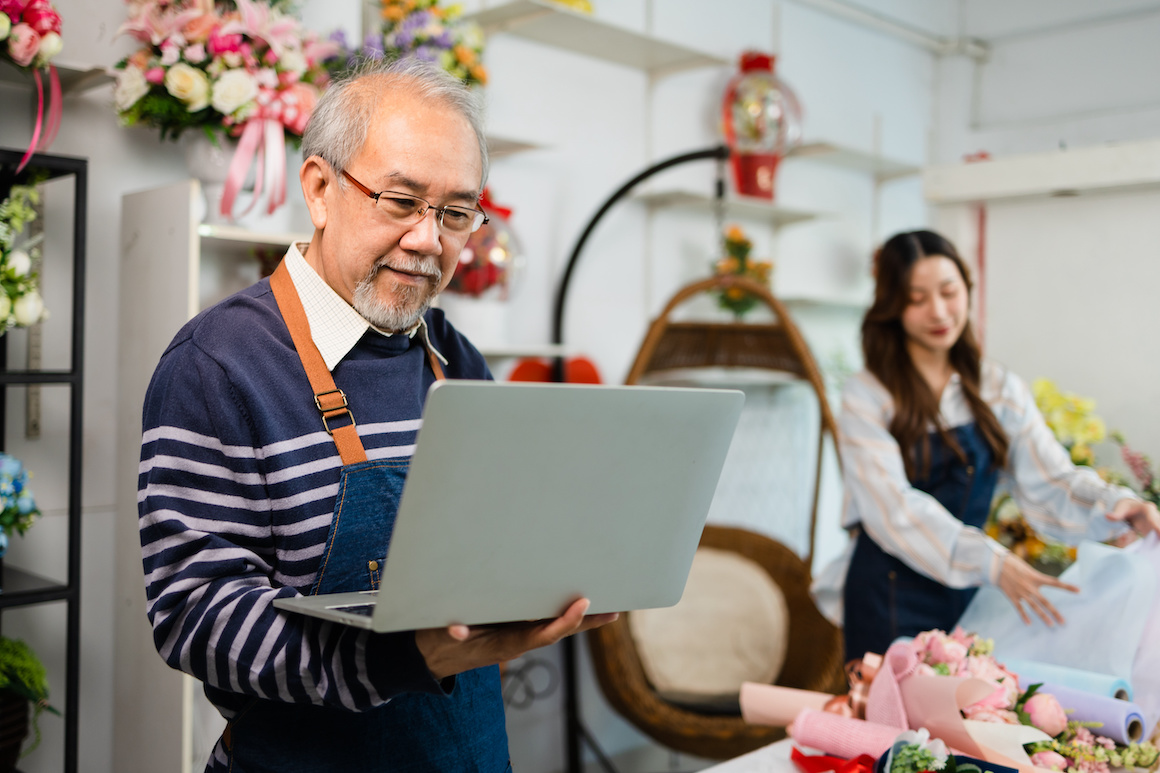 senior man florist talking on smartphone using laptop at florist. portrait of mature male small business owner using laptop and looking at camera in flower shop