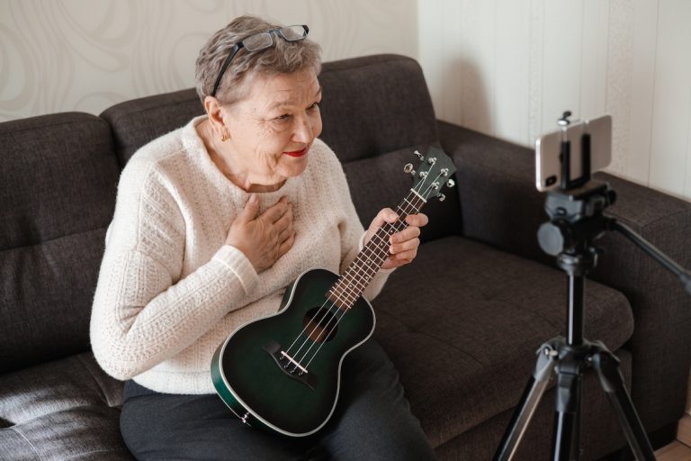 Senior woman with ukulele in her hands, sits on the couch and talks via video chat