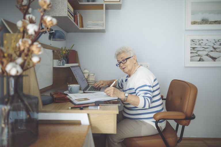 Senior woman at home working on computer writing in notebook