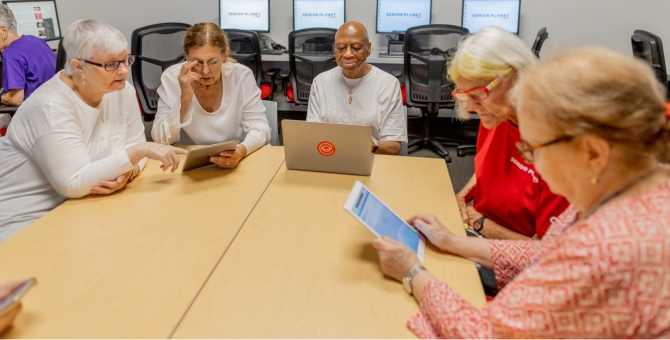 group of older adult women sitting around table using tablets and laptops