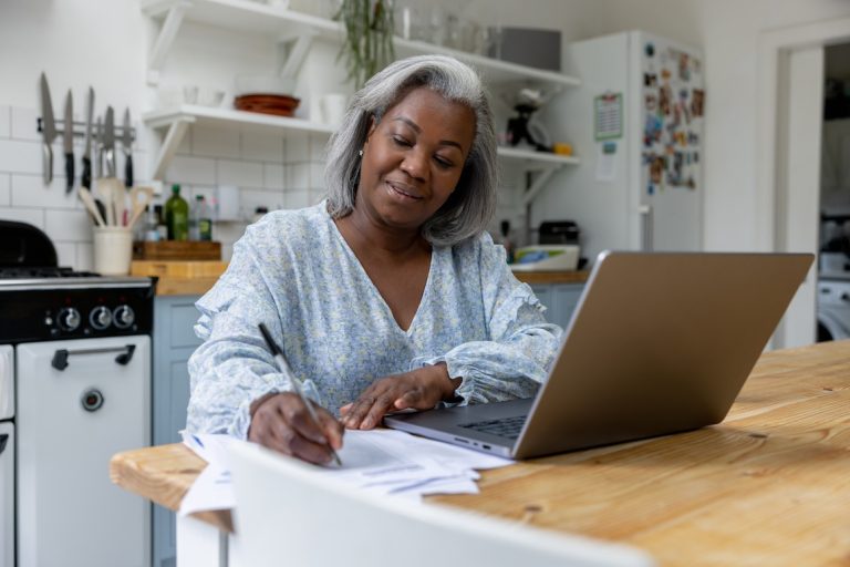 Senior black woman at home writing a letter on paper and using her laptop