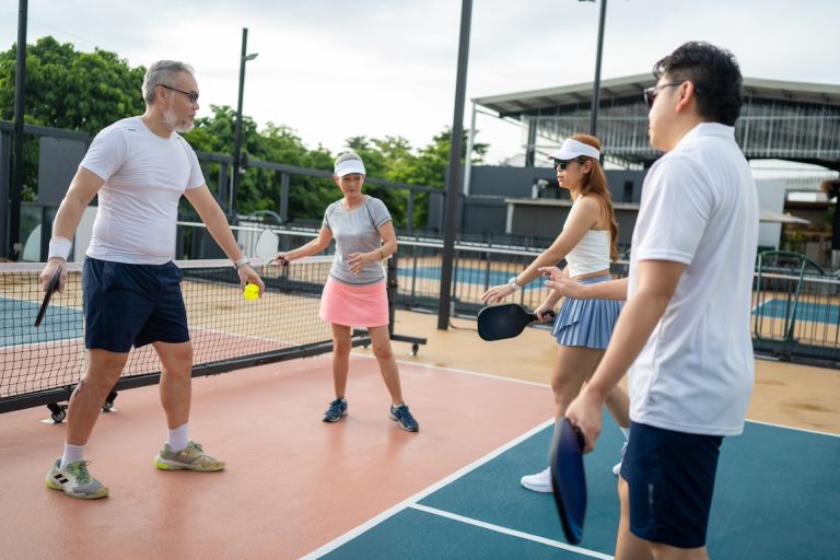 An Asian male pickleball coach is teaching a small group of adult players how to play pickleball on an outdoor court.