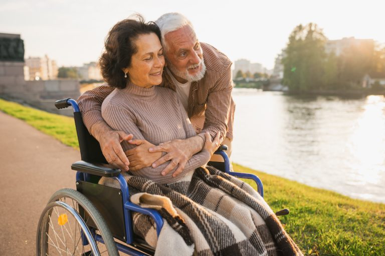 Senior woman in wheelchair walking with caregiver older man on road in park.