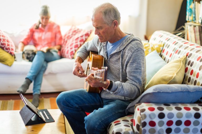 Senior man learning guitar by watching online tutorial. wife reads book in background