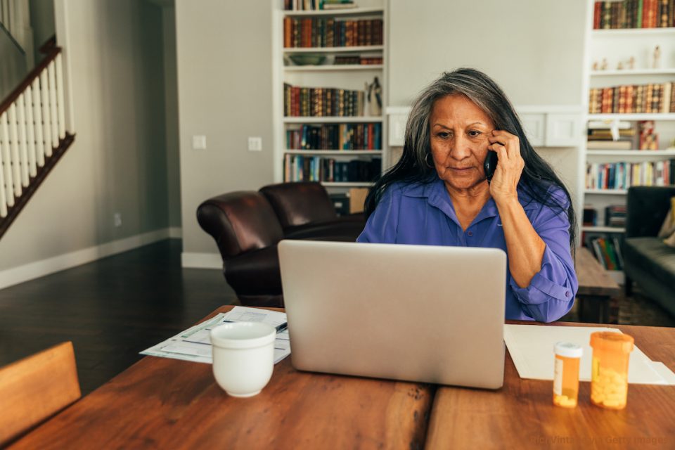 A senior aged woman sits at her kitchen table while paying medical bills, talking with her doctor, and updating medicine prescriptions