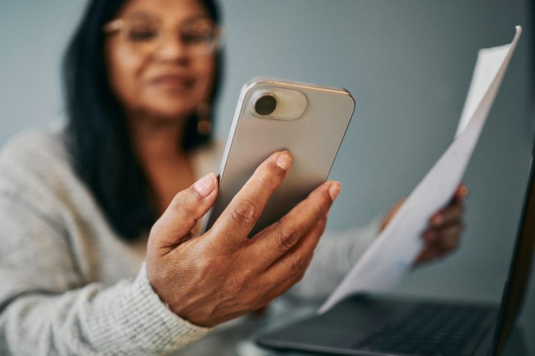 senior woman sits at a desk, reviewing a document and using her mobile phone