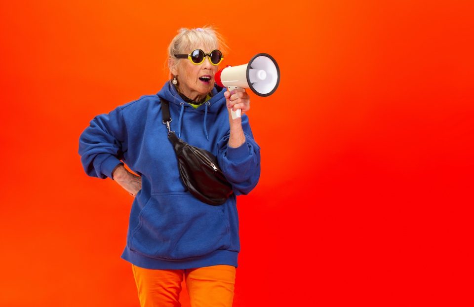 Senior woman in trendy attire shouting in megaphone isolated on bright orange background