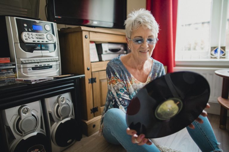 senior woman sitting on the floor in her living room, she is holding a vinyl record in her hands and is listening to music from a record player
