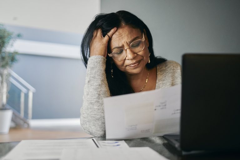 Stressed woman sits at a desk at home, reading a document beside her laptop. She appears worried and stressed, resting her head in her hands while managing her pension finances.