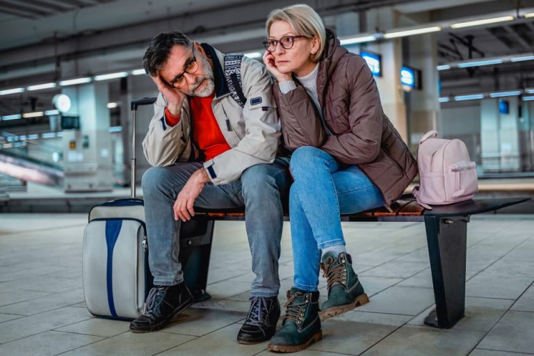 Traveling couple sitting on a bench at a train station showing visible frustration.