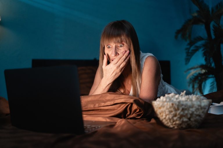 Shocked senior woman eating popcorn and watching horror movie on laptop lying in bed at night.